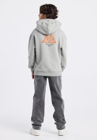 Grijze hoodie met een groot oranje driehoekig logo met de tekst "COLORADO TRAIL CLUB 1969" op de achterkant. Draagt een grijze broek en witte sneakers.