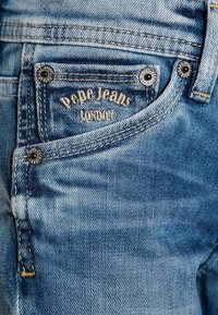 Primer plano de un bolsillo de unos vaqueros azul denim con "Pepe Jeans London" bordado y remaches metálicos en las esquinas del bolsillo.