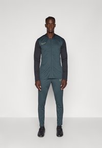 Nike tréningový outfit v tmavom teal a čiernej farbe, obsahujúci zipsový kabát s stojacim golierom a zodpovedajúce zúžené nohavice, doplnené čiernymi športovými topánkami.