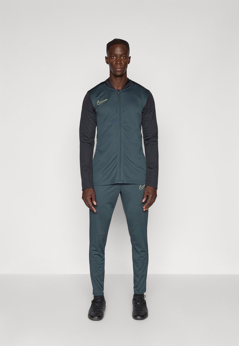 Nike tréningový outfit v tmavom teal a čiernej farbe, obsahujúci zipsový kabát s stojacim golierom a zodpovedajúce zúžené nohavice, doplnené čiernymi športovými topánkami.
