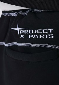 Tela negra con texto bordado en blanco "PROJECT X PARIS" y un logo de estrella, con detalles de costura blanca cerca de la apertura del bolsillo.