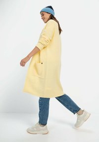 Lange gele cardigan van textuurstof, met een zijzak en ronde zoom. Gecombineerd met blauwe jeans en lichte sneakers.