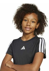 Giovane con capelli ricci che indossa una maglietta nera Adidas con strisce bianche, in piedi con aria sicura e le braccia incrociate, su uno sfondo neutro.