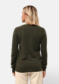 Kvinna med blont hår som bär en mörkgrön långärmad tröja och beige byxor, vänd bort mot en enfärgad ljus bakgrund.