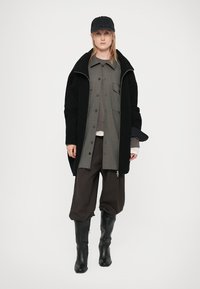 Juodas raukšlėtas paltas virš pilkos marškinių su kišenėmis, derinamas su rudomis laisvomis kelnėmis ir juodais iki kelių batais, užsidėtas juodas kepurė.