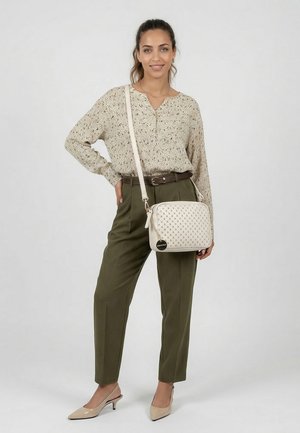 Naine, kes kannab beežikat kudumit ja oliivrohelisi pükse, kandes valget õlakotti mustrilise kujunduse ja kuldse heategijaga.