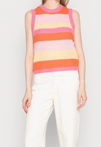 Kvinde iført en ærmeløs strikket top med vandrette orange, pink og gule striber, parret med hvide bukser.
