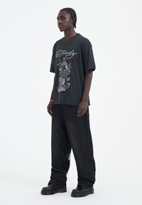 Černé grafické tričko s barevnými vzory, spárované s volnými černými denimovými kalhotami a robustními černými botami.