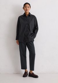 Vrouw staat tegen een witte muur in een losse zwarte button-up blouse, donkere rechte broek en donker bordeauxrode loafers, met één hand in haar zak.