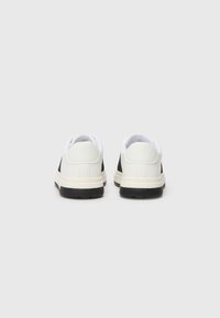 Hvite og svarte sneakers med en glatt tekstur, lavprofil design, gummisåler og minimalistiske detaljer. Bakfra viser de en avrundet hæl.