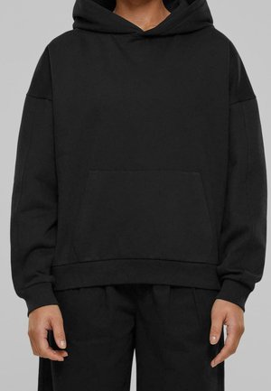 Personne portant un sweat à capuche noir oversize avec une poche kangourou devant et un pantalon noir, debout devant un fond gris uni.