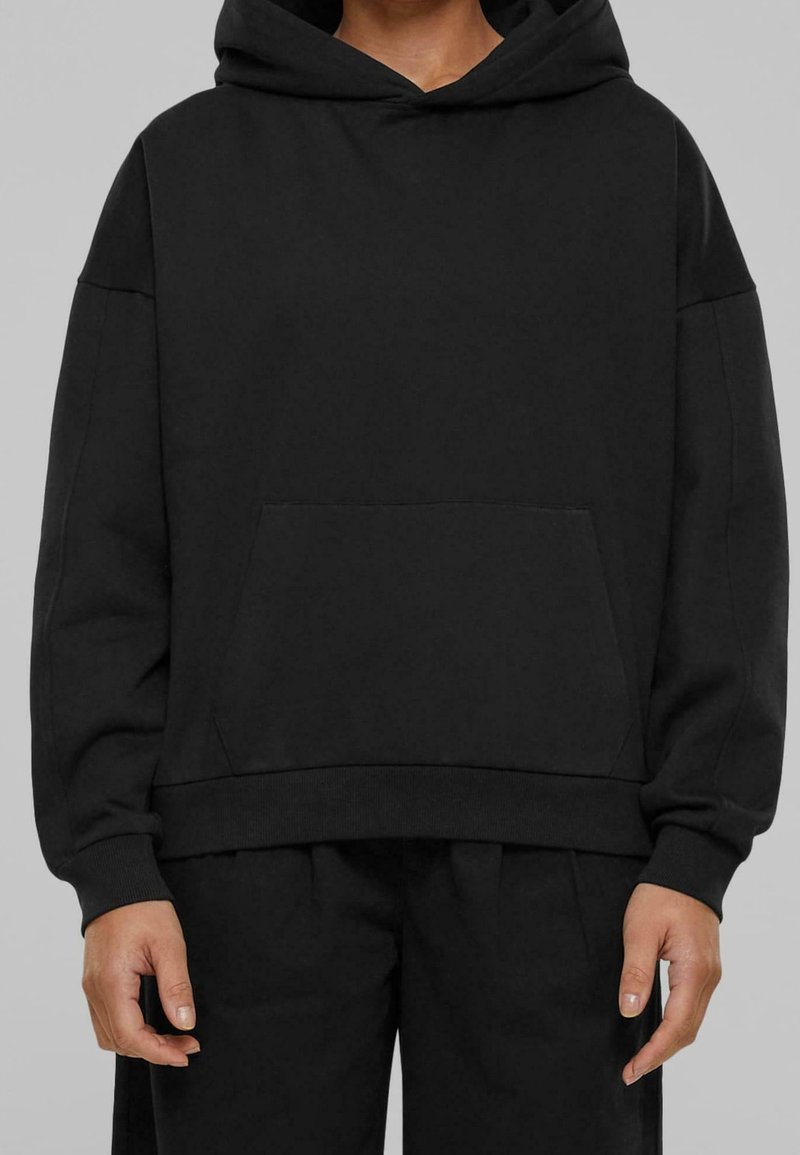 Personne portant un sweat à capuche noir oversize avec une poche kangourou devant et un pantalon noir, debout devant un fond gris uni.
