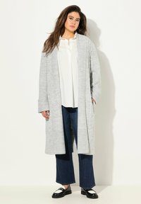 Světle šedý pletený cardigan s žebrovanou texturou a otevřeným předním dílem, kombinovaný s bílou blůzou a tmavými širokými džínami. Černé mokasíny dotvářejí celkový vzhled.