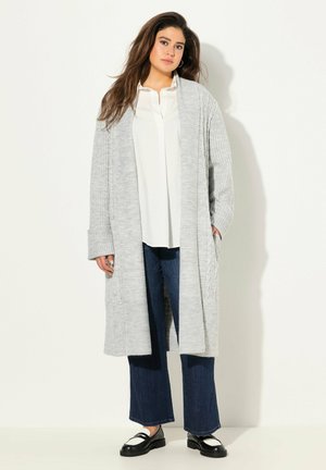 Un cardigan tricoté gris clair avec une texture côtelée et une ouverture à l'avant, associé à une blouse blanche et un jean large foncé. Des mocassins noirs complètent le look.