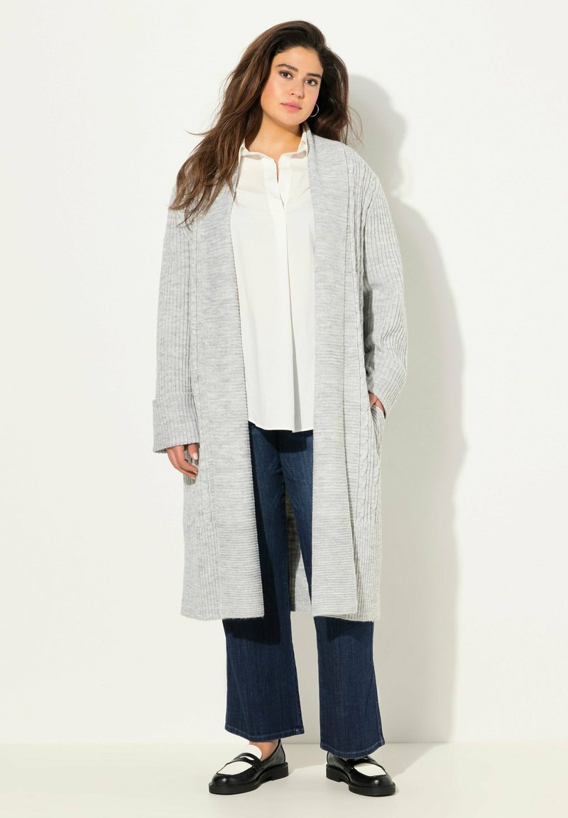 Světle šedý pletený cardigan s žebrovanou texturou a otevřeným předním dílem, kombinovaný s bílou blůzou a tmavými širokými džínami. Černé mokasíny dotvářejí celkový vzhled.