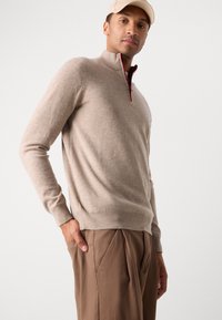 Beige knitted pullover with a zippered collar, featuring a red accent. Soft texture and fitted design paired with brown trousers.