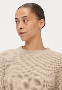 Beige gebreide trui met een textuurpatroon en een ronde hals. Model heeft een eenvoudige hoopoorbel, haar strak naar achteren gekamd en een neutrale uitdrukking.
