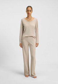 Vrouw met gevlochten haar, gekleed in een beige langemouwen shirt en wijde broek, blootsvoets staand voor een effen lichte achtergrond.