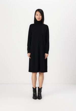 Robe noire à manches longues avec un col haut, présentant une silhouette ample qui tombe jusqu'au mollet. Portée avec des bottes noires à la cheville.