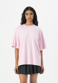 T-shirt oversize rosa chiaro realizzata in tessuto morbido, con maniche corte e scollatura rotonda. Abbinata a una gonna plissettata scura.