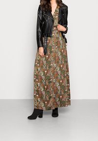 Zwarte leren cropped jacket over een lange, gemusterde, groen- en bruinbloemige maxi-jurk. Gecombineerd met zwarte hiel enkellaarzen.