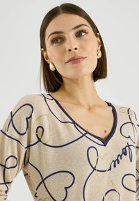 Top de punto beige con patrón de corazones en azul oscuro y detalles dorados. Escote en V con acabado texturizado. Modelo lleva pendientes grandes dorados.