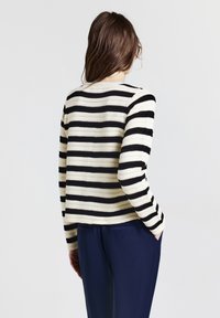 Knitted sweater in cream with bold black horizontal stripes, featuring a round neckline and long sleeves. Worn with dark blue trousers.