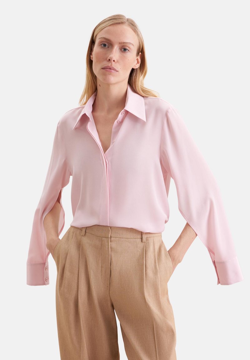 Femme portant un chemisier rose clair avec des fentes aux manches et un pantalon plissé beige taille haute, debout les mains dans les poches.