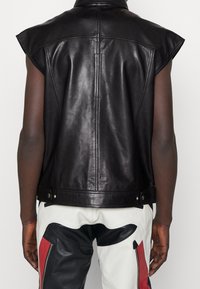 Svart läderväst med breda axlar och synliga sömmar. Har tryckknappar vid midjan, tillsammans med krämfärgade och svarta byxor.