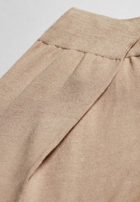 Beige stof med en glat tekstur, der har en bred linning og subtile plisseringer nær taljelinjen, orienteret vandret.