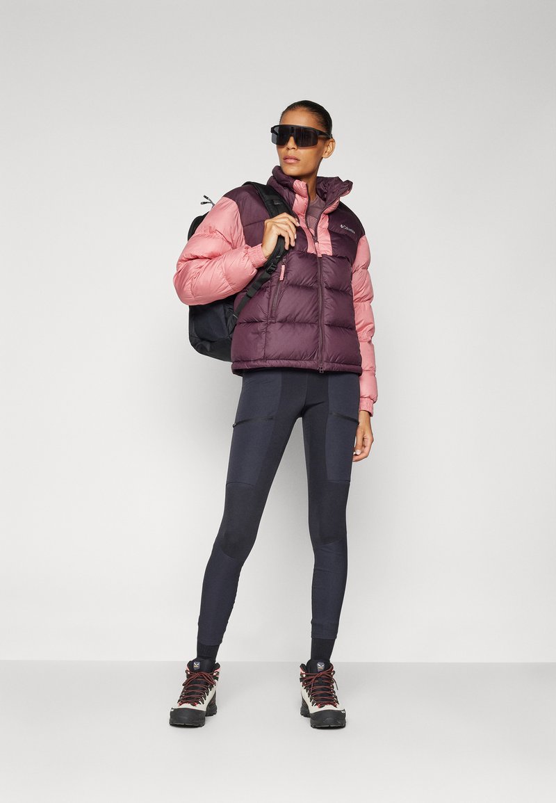 Gilet imbottito rosa e viola scuro, abbinato a leggings neri elasticizzati. Il modello indossa stivali da trekking neri e occhiali da sole, portando uno zaino nero.