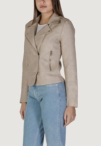 Giacca da motociclista in similpelle beige con colletto a punte, zip asimmetrica e due tasche anteriori con zip. Abbinata a jeans in denim azzurri chiari.