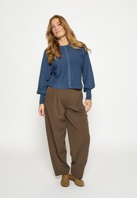 Blauw geribd blouse met knopen en ballonmouwen, gecombineerd met bruine geplooide broek. Model draagt lichtbruine schoenen en staat tegen een neutrale achtergrond.
