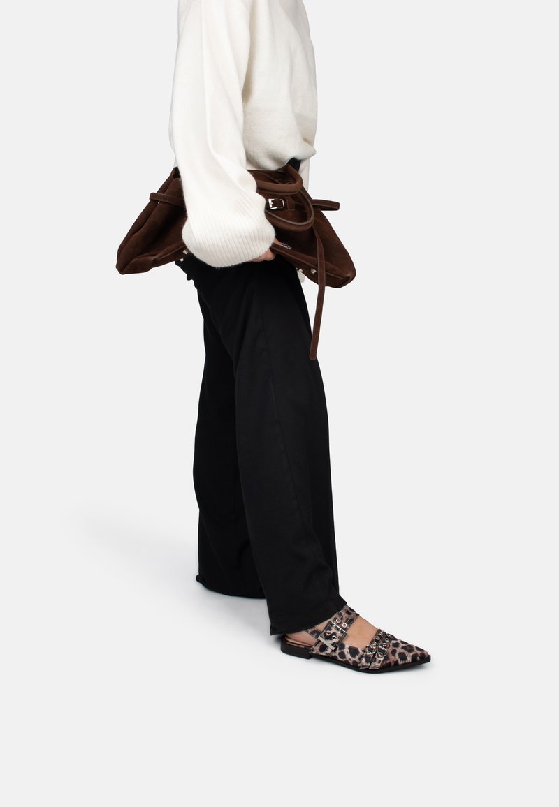 Persoon in zwarte broek en witte trui houdt een grote bruine suède handtas vast, draagt puntige leopardprint flats met gespen en kettingen.