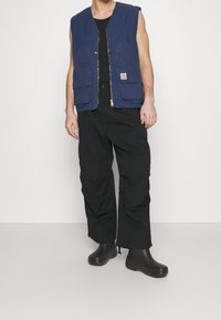 Niebieska bezrękawnik z zamkiem błyskawicznym z przodu, dwoma przednimi kieszeniami i żółtymi akcentami, połączony z czarnymi spodniami cargo i czarnym obuwiem.