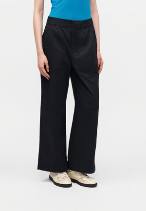 Άτομο που φοράει μαύρο παντελόνι με φαρδιές φάσες και εμπρός τσέπες, συνδυασμένο με μπεζ αθλητικά παπούτσια και μπλε τοπ, στέκεται μπροστά σε μονόχρωμο φόντο.