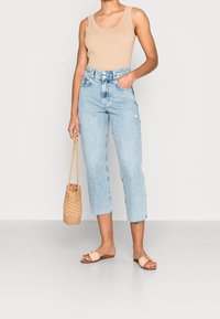 Žena oblečená v béžovém tílku, světle modrých zkrácených džínách, béžových pletených šlapkách, držící béžovou pletenou kabelku po boku.