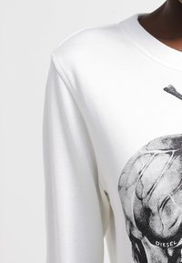 Vit sweatshirt med en grafisk tryck av ett dödskallemotiv på framsidan, rund hals och textil med struktur. Sidsömmar synliga.