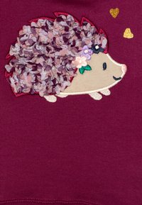 Vêtement marron avec un hérisson brodé et une texture florale superposée au dos, agrémenté de petits cœurs et d'accents floraux.