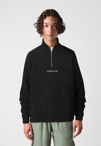 Musta fleece-neule korkealla kauluksella ja neljänneksen vetoketjulla, jossa on etupuolella tasku ja "Originals Studio" kirjailtuna valkoisena.
