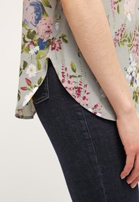 Haut à manches courtes à motif floral en tissu gris clair, avec des fleurs roses, vertes et bleues. Assorti à un jean en denim bleu foncé avec poches.