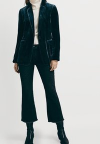 Femme portant un blazer en velours sombre, un pull col roulé crème, un pantalon évasé noir raccourci et des bottines noires, debout devant un fond uni.