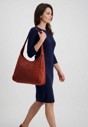 Femme en robe bleu marine marchant, portant un grand sac à bandoulière en cuir marron sur son épaule droite sur fond uni.