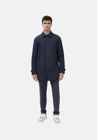 Cappotto navy con tasche laterali, colletto e silhouette dritta, abbinato a pantaloni scuri e sneakers bianche su uno sfondo semplice.