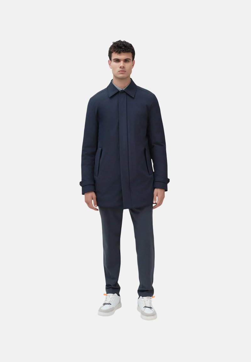 Cappotto navy con tasche laterali, colletto e silhouette dritta, abbinato a pantaloni scuri e sneakers bianche su uno sfondo semplice.
