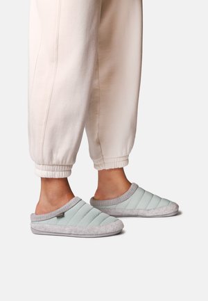 Person iført lysegrå, quiltede slip-on tøfler og cremefarvede svedbukser med manchetter, stående på en ensfarvet lys baggrund.