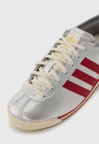 Srebrne usnjene športne čevlje s smetanastimi vezalkami. Imajo tri rdeče črte in retro dizajn. Vključujejo zlat logo na jeziku.