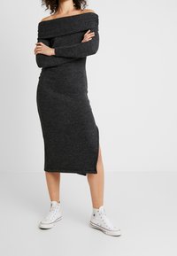 Robe noir gris à épaules dénudées avec des manches longues et une fente sur le côté, fabriquée en tissu tricoté texturé, associée à des baskets montantes blanches.
