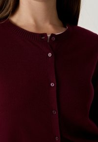 Gilet bordeaux en tissu doux, avec un col rond, une bordure côtelée et de de petits boutons violets le long de la patte de boutonnage.