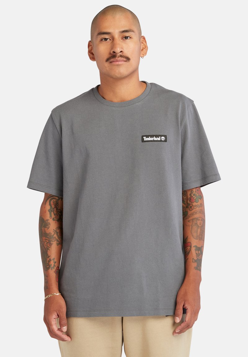 T-shirt grigia in cotone con collo rotondo e maniche corte, caratterizzata da un piccolo patch del logo nero Timberland sull'area del petto sinistro.